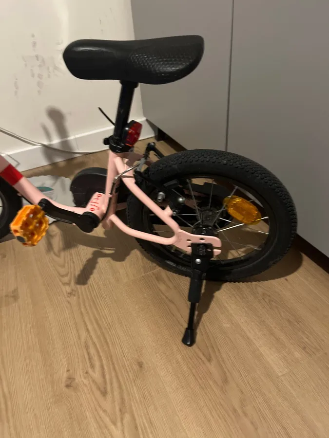 A pink 14 inch bike resting on its newly fitted kickstand indoors
