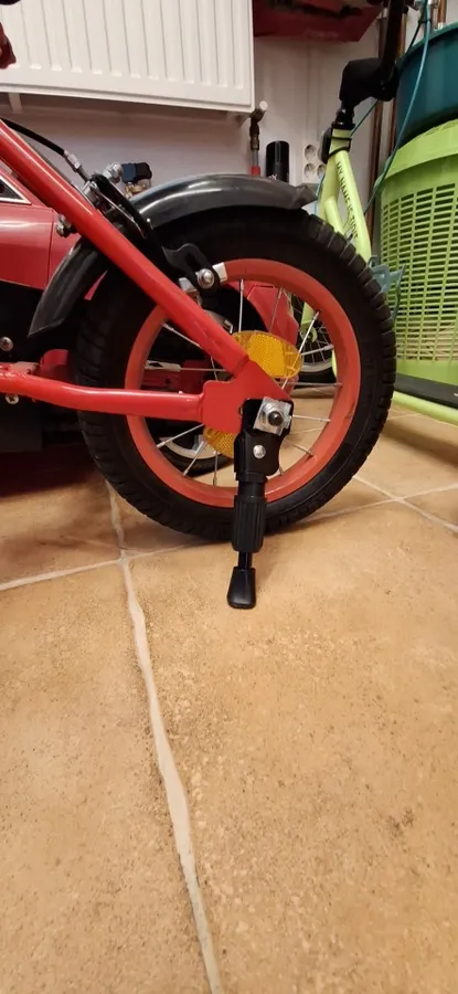 Kickstand holding up a red 12-inch kids bike on a tiled floor, photographed by a verified buyer